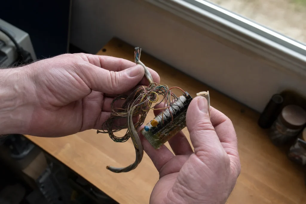 Händer som undersöker en trasslig telefonkabel och ett litet kretskort vid ett fönster i en hemmaverkstad.