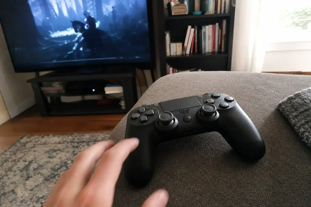 En hand sträcker sig mot en spelkontroll på soffan, med pausat spel i bakgrunden, apropå att EA tillhör Saudiarabien och J...